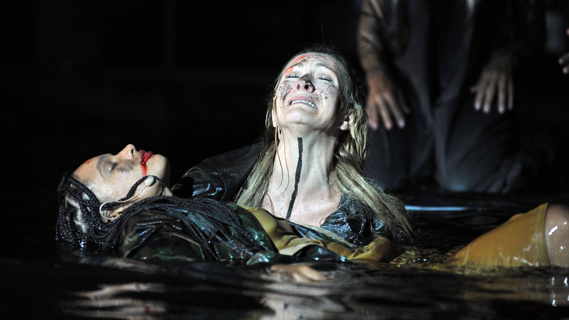 Foto von der Inszenierung: Eine weinende Frau hält eine anscheind tote Frau mit den Armen über Wasser.