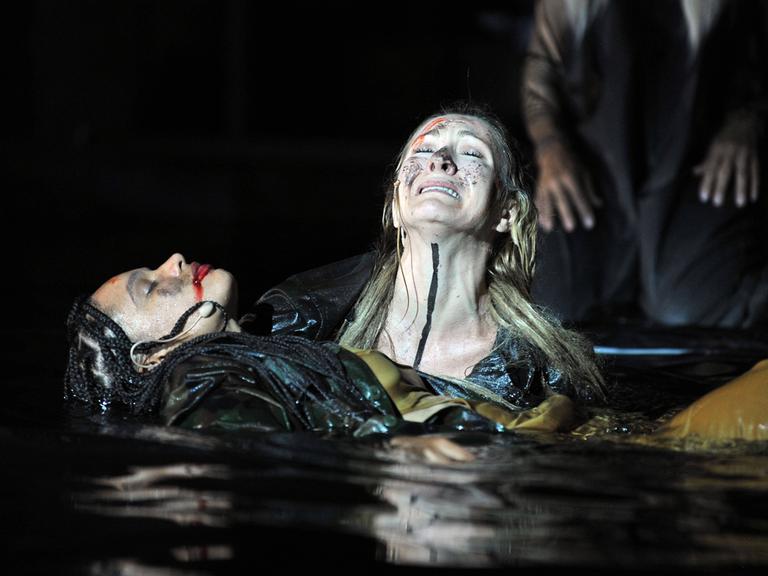 Foto von der Inszenierung: Eine weinende Frau hält eine anscheind tote Frau mit den Armen über Wasser.