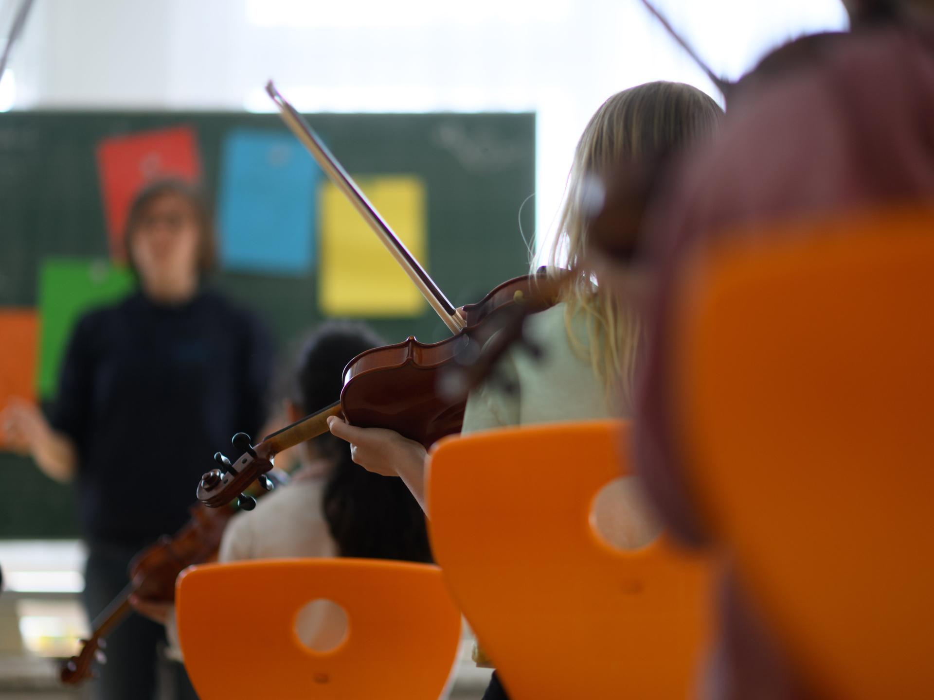 In einer Schulklasse sitzt ein Mädchen mit einer Geige auf einem Stuhl, im Hintergrund sieht man eine Tafel und eine Lehrerin.