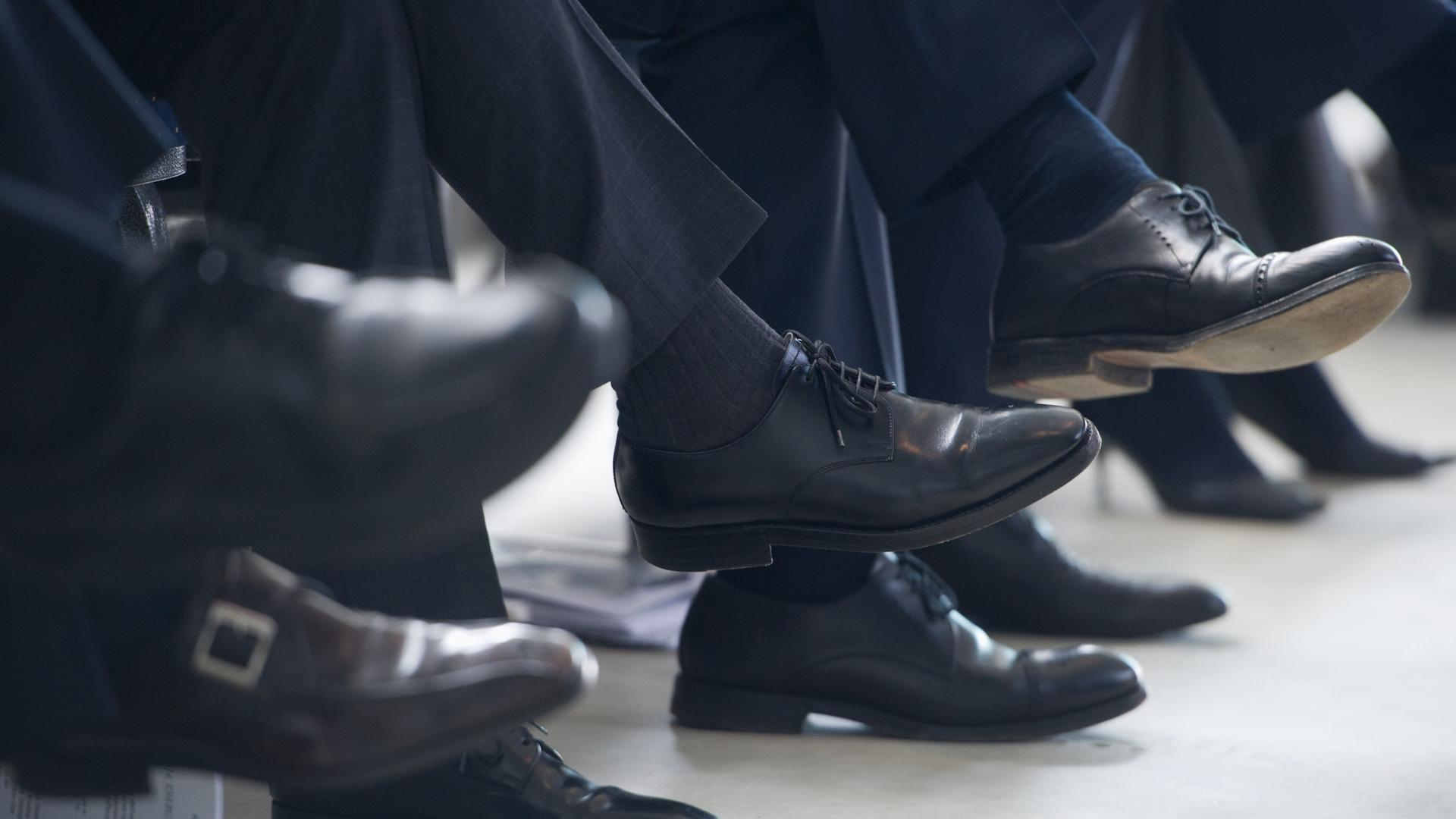 Männer in Anzugshosen sitzen nebeneinander; das Foto zeigt ihre Schuhe; es sind schwarze Schuhe.