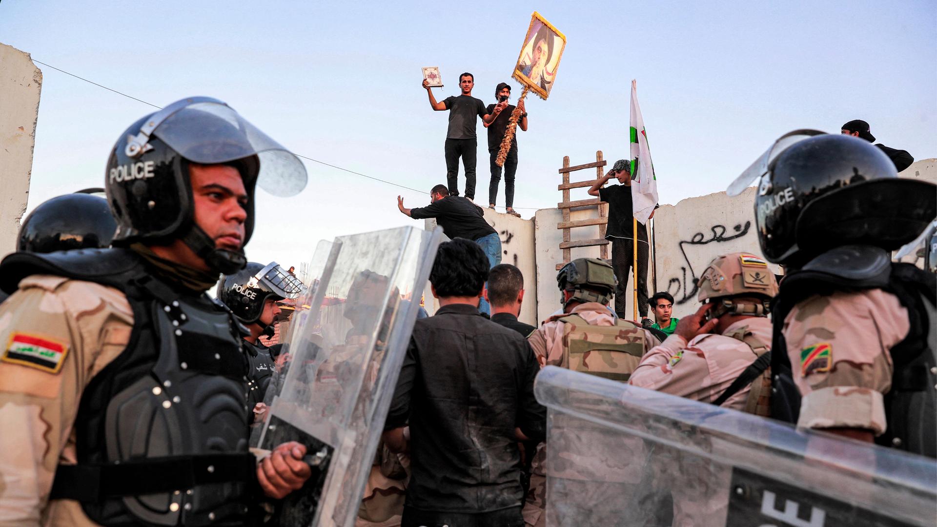Protestierende Männer mit einem Koran stehen auf dem Zaun der schwedischen Botschaft in Bagdad. Uniformierte Polizeibeamte versuchen die Menschen zu zerstreuen.