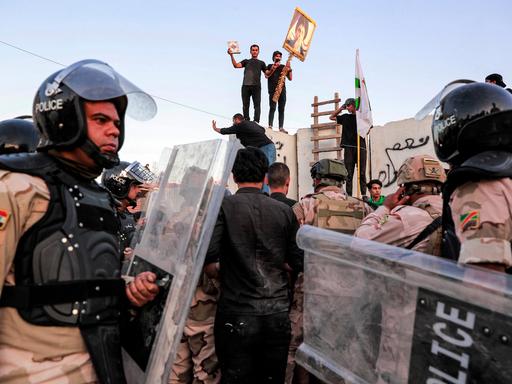 Protestierende Männer mit einem Koran stehen auf dem Zaun der schwedischen Botschaft in Bagdad. Uniformierte Polizeibeamte versuchen die Menschen zu zerstreuen.