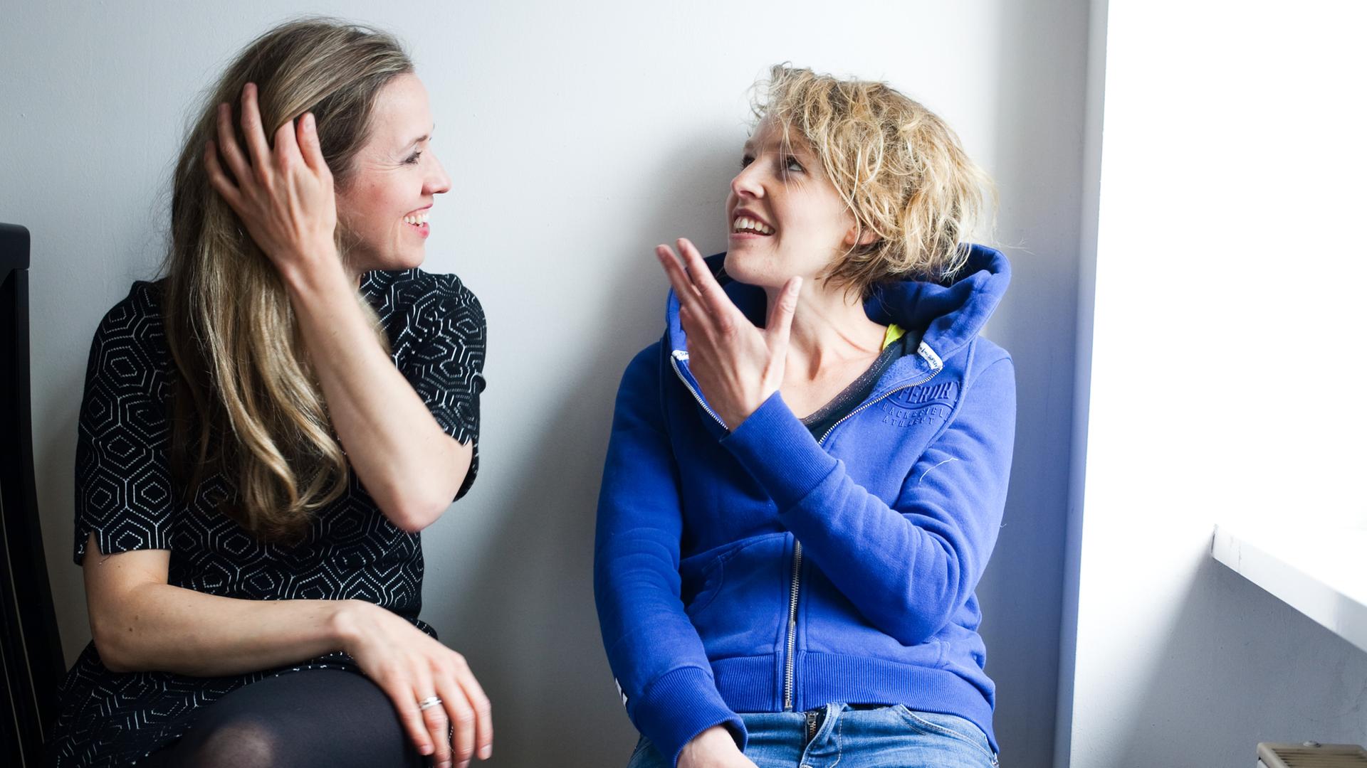 Zwei junge Frauen sitzen vor einer Wand und unterhalten sich. Die Frau links im Bild, Annika Sophie Mendrala, hört lächelnd zu. Verena Usemann, die Frau rechts, gestikuliert beim Sprechen und schaut leicht nach oben. 
