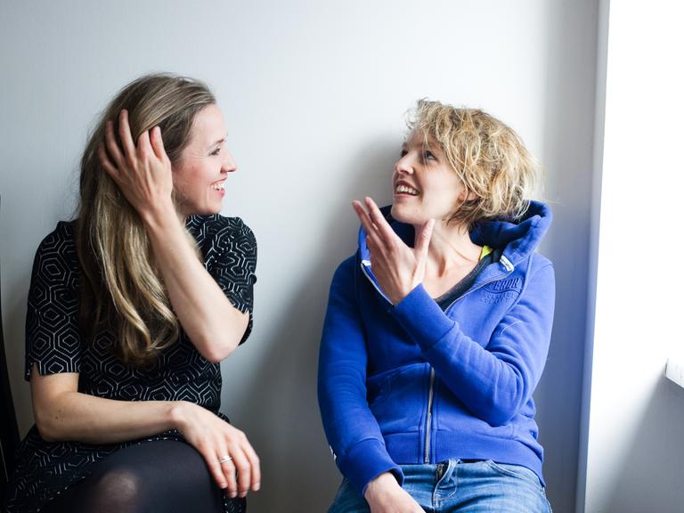 Zwei junge Frauen sitzen vor einer Wand und unterhalten sich. Die Frau links im Bild, Annika Sophie Mendrala, hört lächelnd zu. Verena Usemann, die Frau rechts, gestikuliert beim Sprechen und schaut leicht nach oben. 