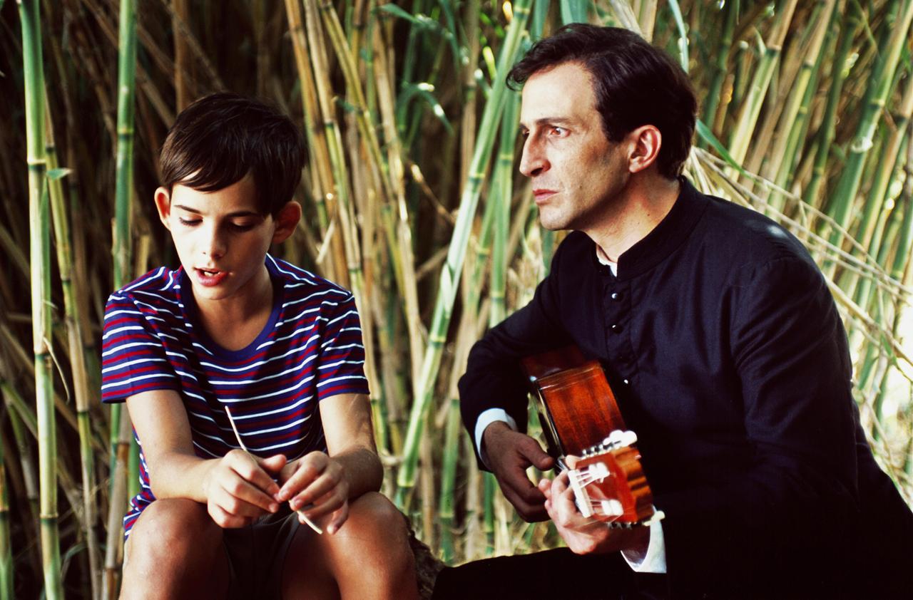 Szene aus dem Film "Schlechte Erziehung" - ein Junge sitzt mit einem Pfarrer im Gras, der Pfarrer spielt Gitarre