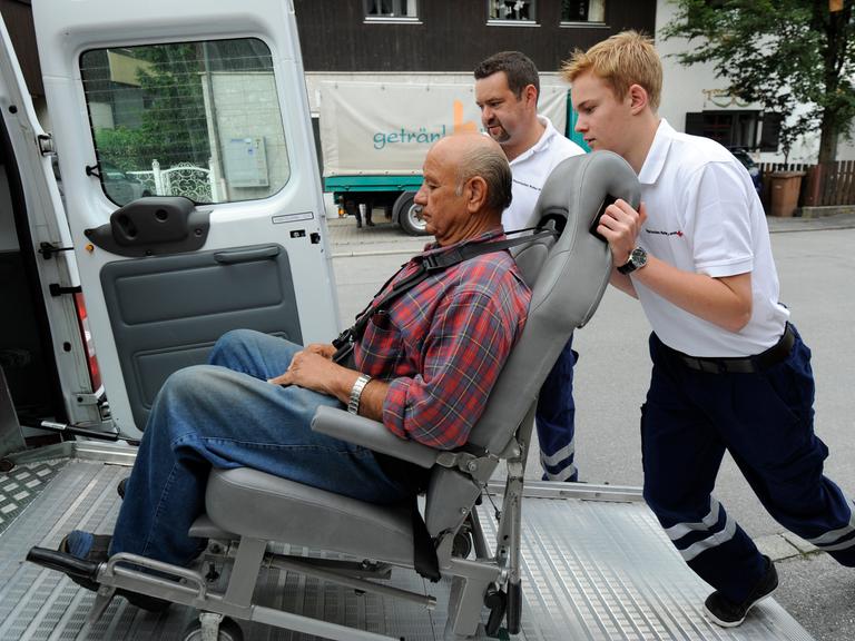 Ein junger Mann schiebt einen Rollstuhl mit einem Mann darauf in einen Transporter 