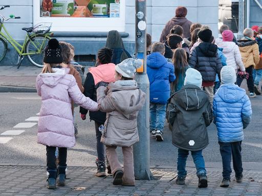 Kinder eines Kindergartens sind von hinten zu sehen, wie sie Hand in Hand die Straße überqueren. 