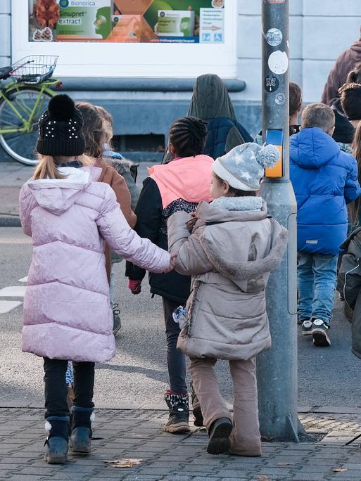 Kinder eines Kindergartens sind von hinten zu sehen, wie sie Hand in Hand die Straße überqueren. 