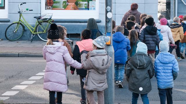 Kinder eines Kindergartens sind von hinten zu sehen, wie sie Hand in Hand die Straße überqueren. 