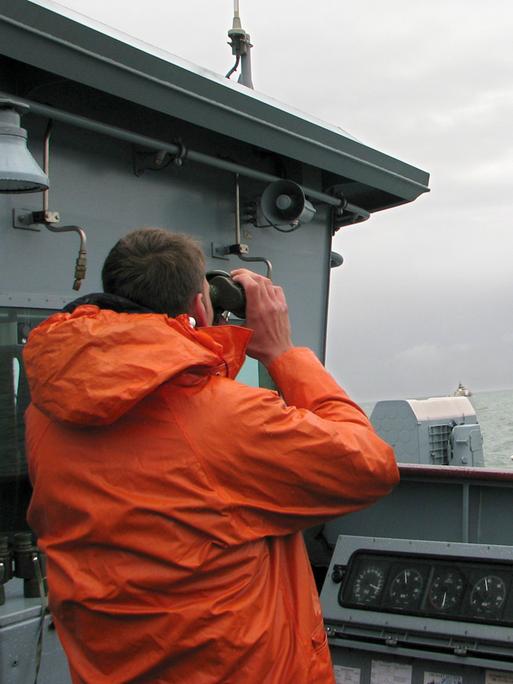 Ein Marinesoldat in Regenjacke steht als Ausguck auf einem Schiff und betrachtet das Meer durch ein Fernglas.