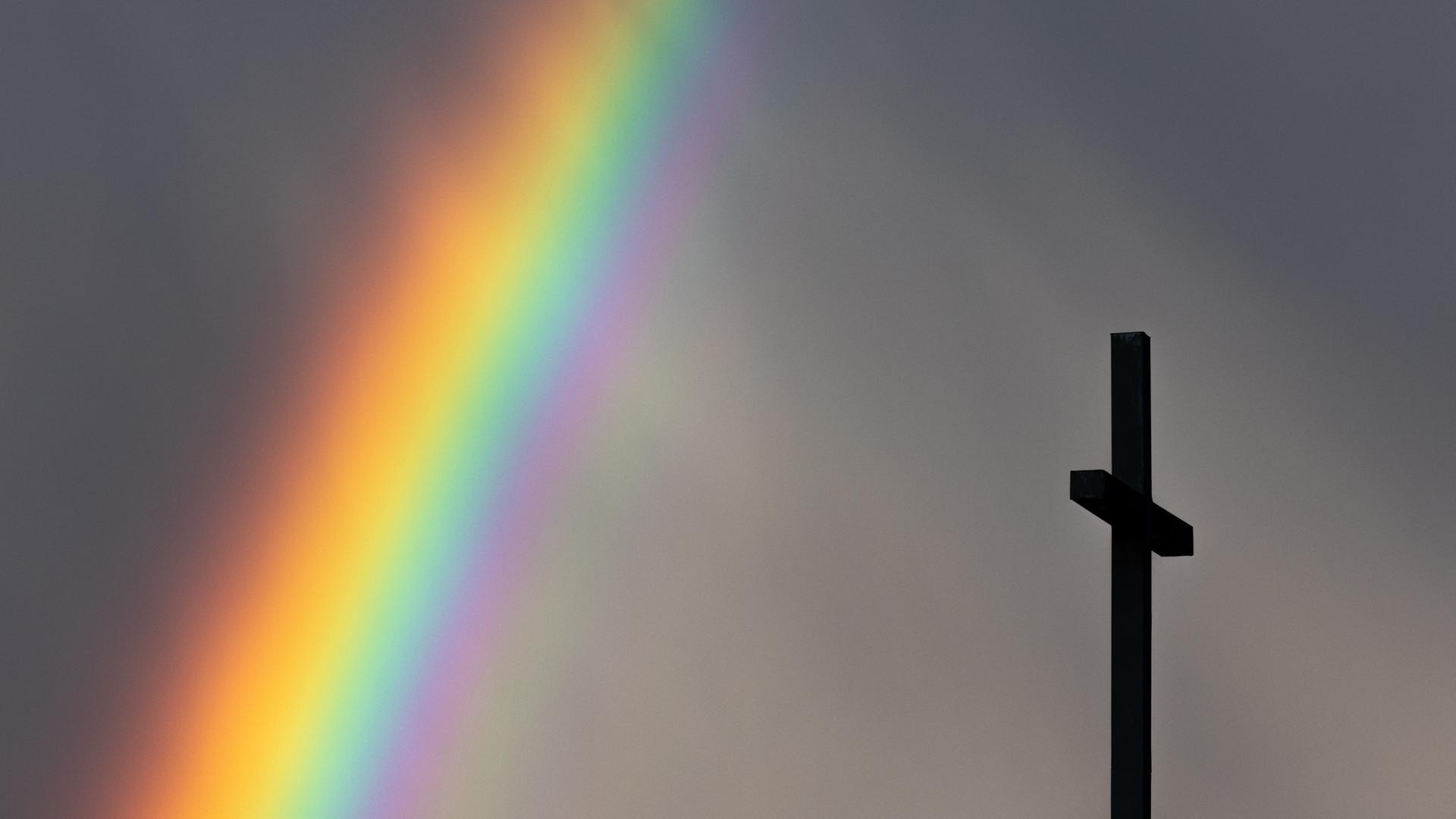Ein kräftiger Regenbogen ist am dunklen Himmel neben dem Kreuz einer Kirche zu sehen.