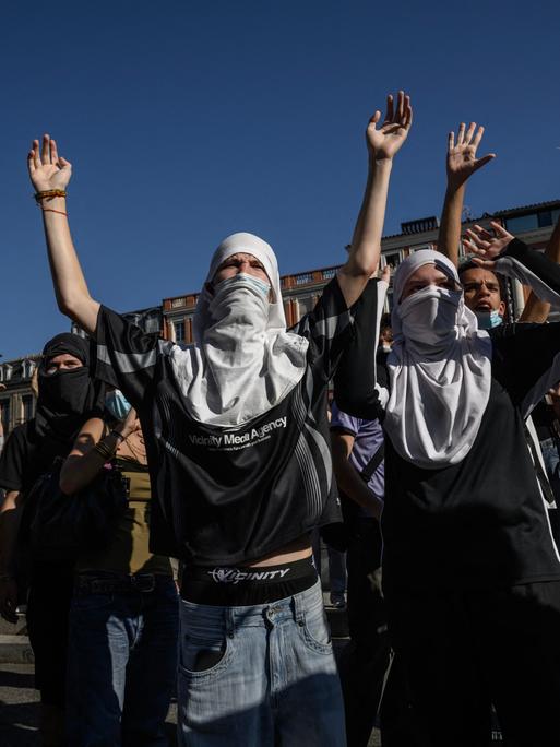 Teilweise vermummte Personen stehen auf einer Demonstration und heben ihre Hände in die Luft