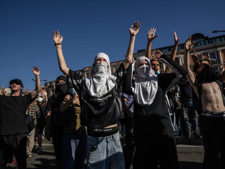 Teilweise vermummte Personen stehen auf einer Demonstration und heben ihre Hände in die Luft