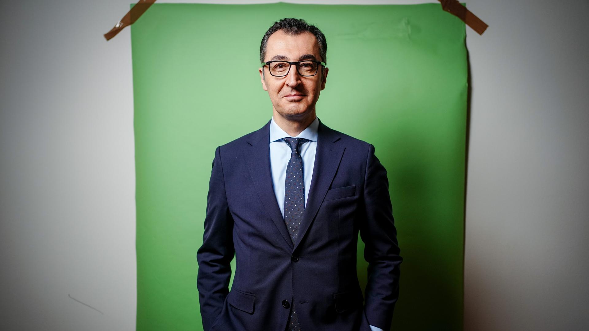 Cem Özdemir steht vor einem an die Wand geklepten grünen Hintergrund. Er trägt einen dunkelblauen Anzug und Krawatte.