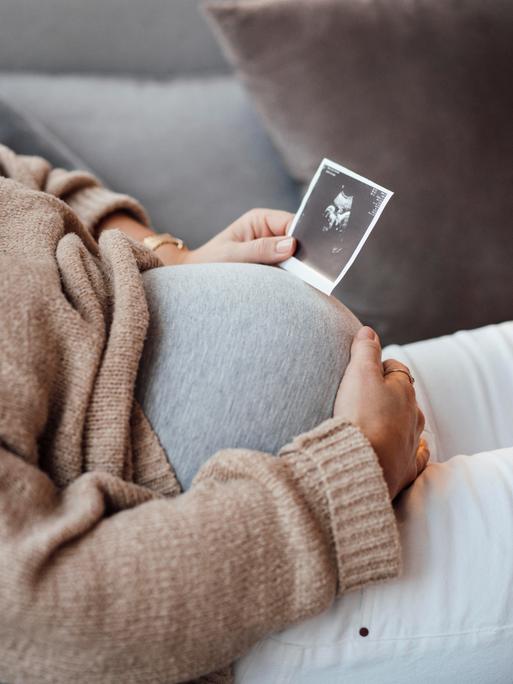 Eine Schwangere sitzt auf einem Sofa, vor ihren Bauch hält sie eine Ultraschallaufnahme.