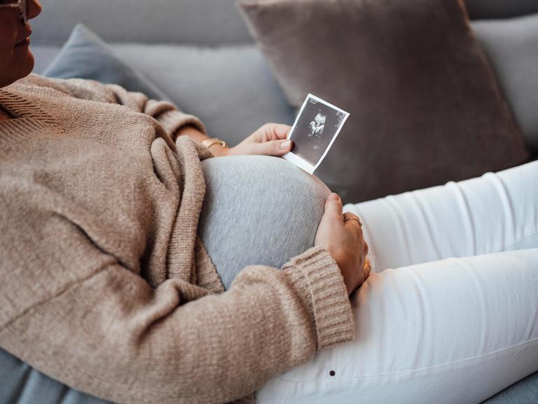 Eine Schwangere sitzt auf einem Sofa, vor ihren Bauch hält sie eine Ultraschallaufnahme.