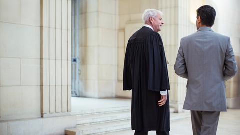 Ein Richter und ein Mann im Anzug laufen an einem Gerichtsgebäude vorbei.