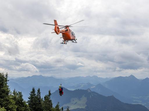 Ein Rettungshubschrauber in den Alpen, der an einem Seil zwei Personen transportiert.
