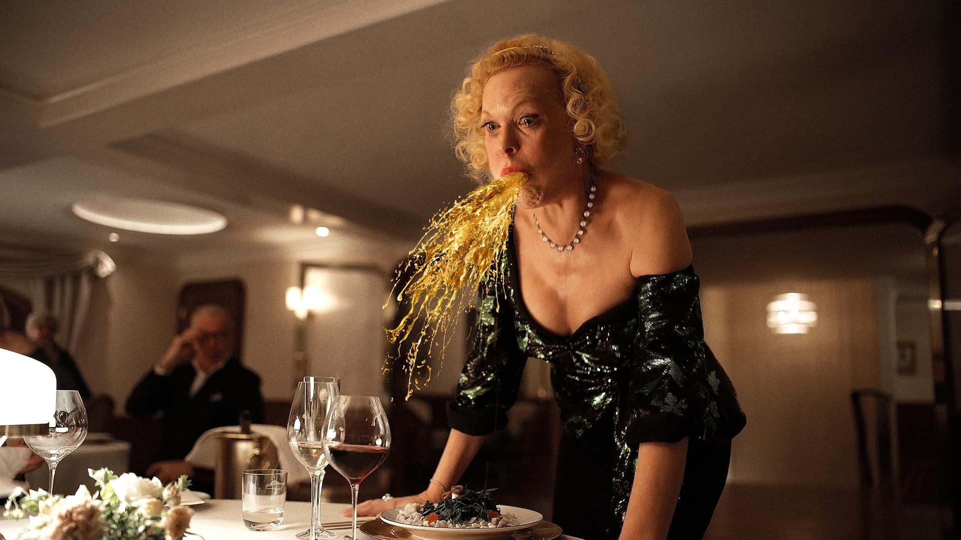 Filmszene aus "Triangle of Sadness": Eine Frau speiht eine Art Goldregen an einem festlichen gedeckten Tisch.