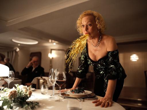 Filmszene aus "Triangle of Sadness": Eine Frau speiht eine Art Goldregen an einem festlichen gedeckten Tisch.