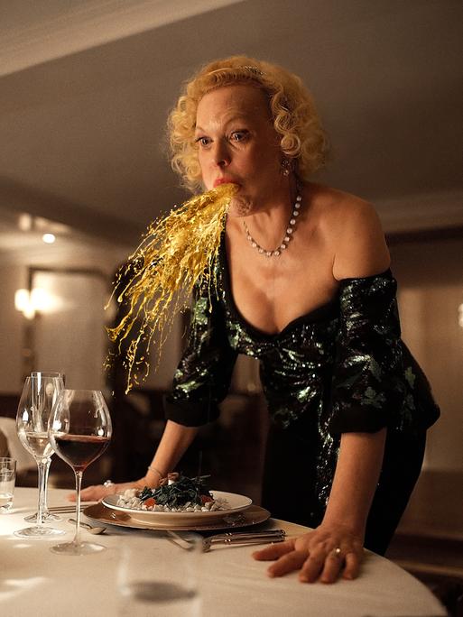 Filmszene aus "Triangle of Sadness": Eine Frau speiht eine Art Goldregen an einem festlichen gedeckten Tisch.
