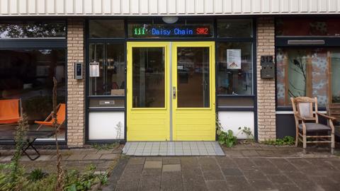 Eine leuchtend gelbe Eingangstür in einer Hausfront mit großen Fenstern und Leuchtschrift über der Tür