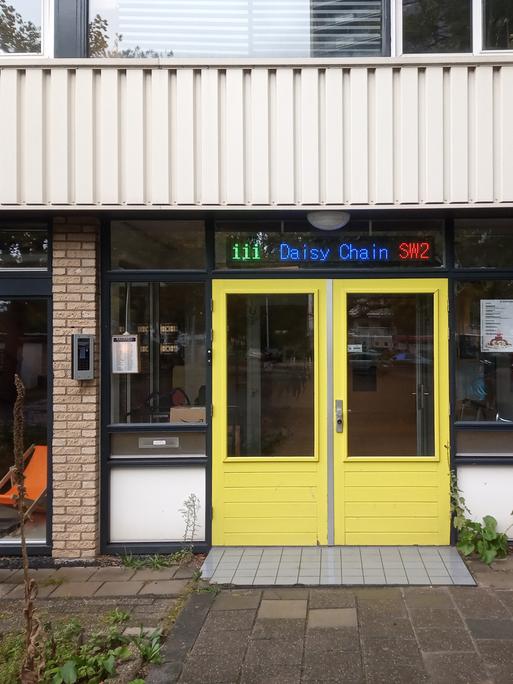 Eine leuchtend gelbe Eingangstür in einer Hausfront mit großen Fenstern und Leuchtschrift über der Tür