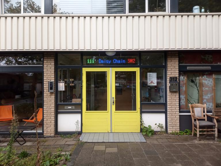 Eine leuchtend gelbe Eingangstür in einer Hausfront mit großen Fenstern und Leuchtschrift über der Tür