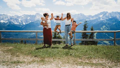 Zwei Mütter posieren mit ihren Kindern vor einer Berglandschaft, zwei Kinder springen, ein kleines ist auf dem Arm