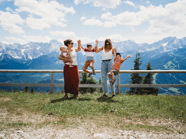 Zwei Mütter posieren mit ihren Kindern vor einer Berglandschaft, zwei Kinder springen, ein kleines ist auf dem Arm