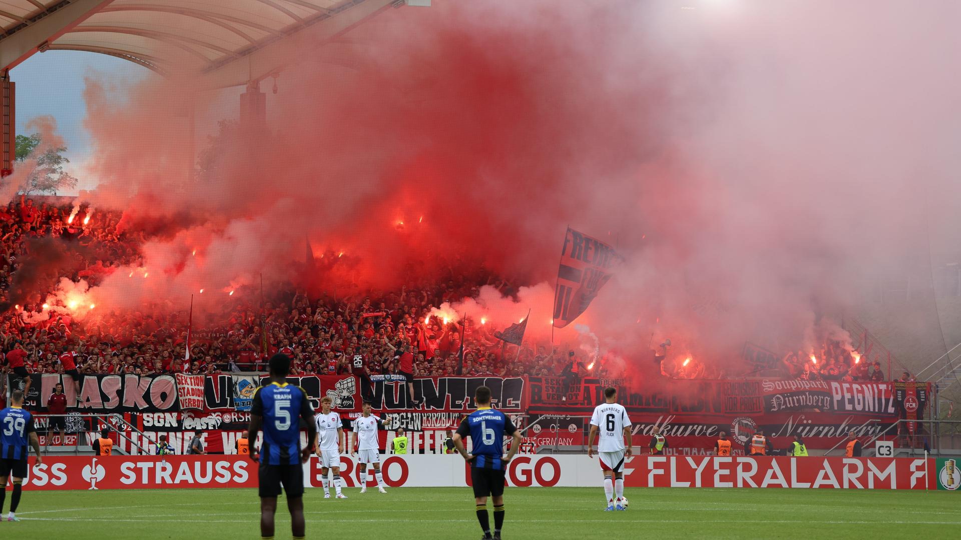 Auf dem Rasen mehrere Spieler von Saarbrücken und Nürnberg - dahinter ist ein in roten Rauch gehüllter Fanblock zu sehen.