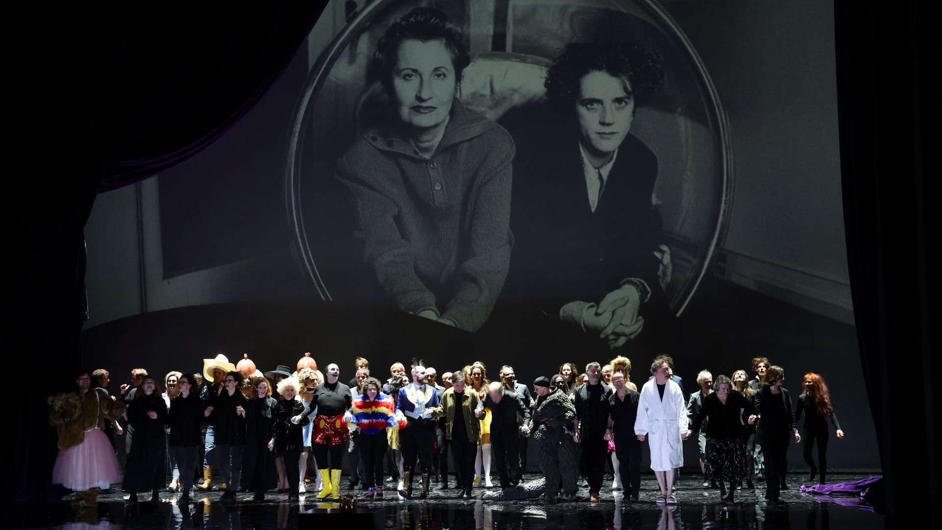 Ensemble und Regieteam von "Monster's Paradise" beim Schlussapplaus der Uraufführung in Hamburg, über ihnen Bilder von Elfriede Jelinek (links) und Olga Neuwirth