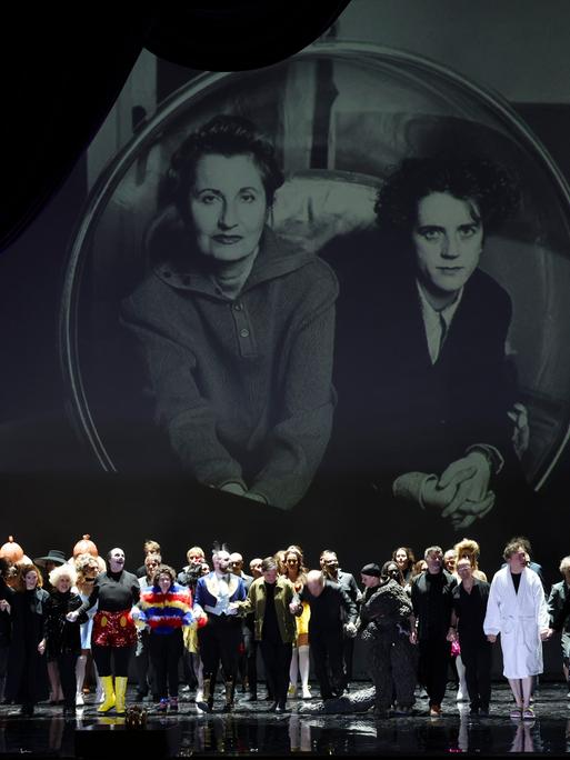 Ensemble und Regieteam von "Monster's Paradise" beim Schlussapplaus der Uraufführung in Hamburg, über ihnen Bilder von Elfriede Jelinek (links) und Olga Neuwirth