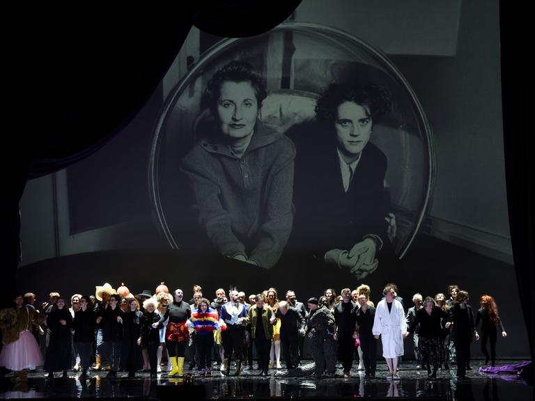 Ensemble und Regieteam von "Monster's Paradise" beim Schlussapplaus der Uraufführung in Hamburg, über ihnen Bilder von Elfriede Jelinek (links) und Olga Neuwirth