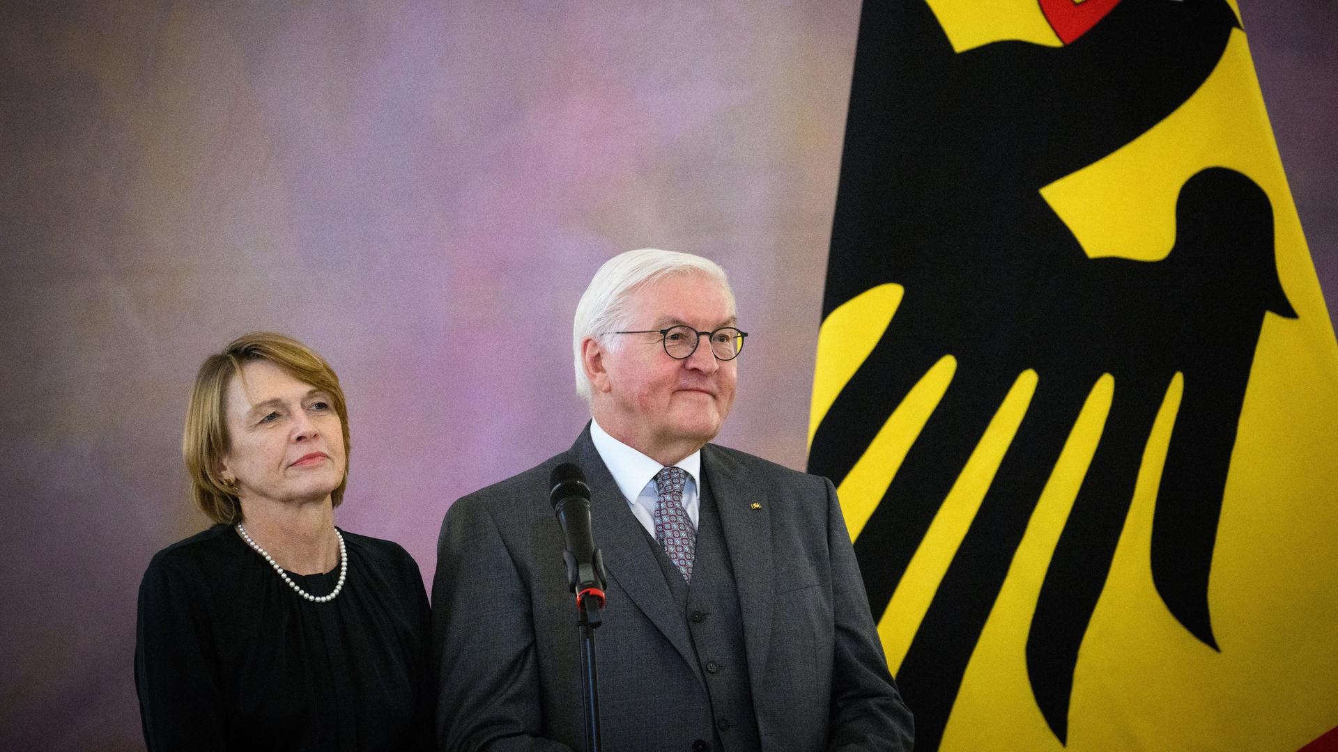 Die beiden stehen lächelnd nebeneinander vor einer lila-grauen Wand. Neben ihnen die gelbe Flagge mit dem schwarzen Bundesadler.