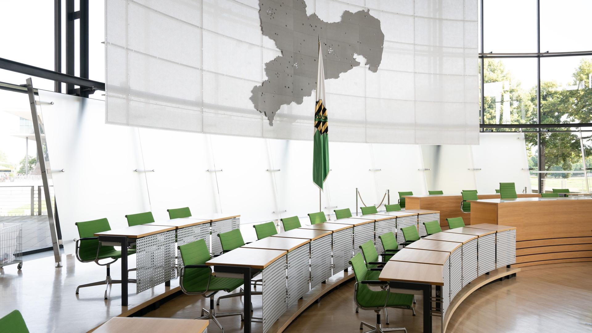 Leere Stühle an der Regierungsbank im Plenarsaal des Landtag. Dahinter steht die sächsische Landesflagge und ein Umriss des Bundeslandes an der Wand.