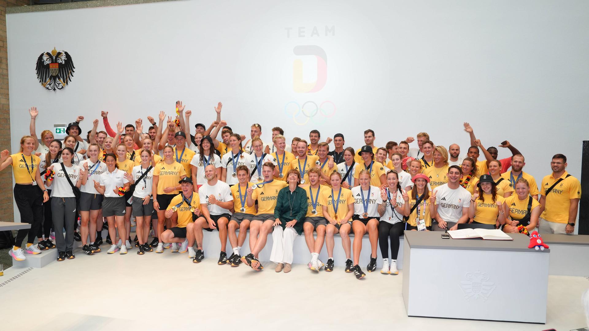 Viele deutsche Olympiateilnehmer in gelben oder weißen Shirts auf einem Gruppenbild. In der Mitte die Kölner Oberbürgermeisterin Reker. Sie Sportler lachen und halten ihre Medaillen hoch. 