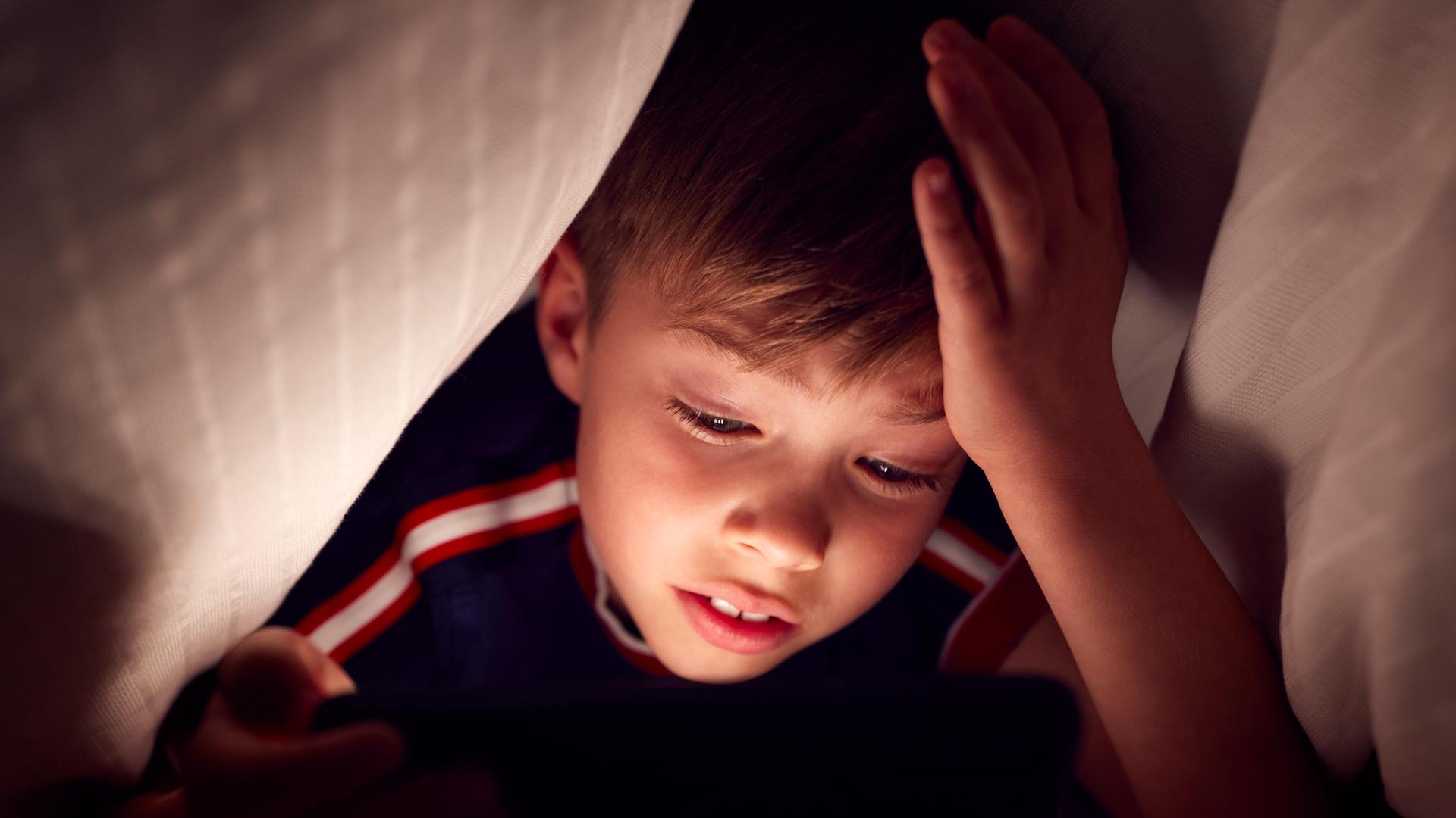 Ein Junge schaut unter der Bettdecke auf sein Tablet oder Smartphone.
