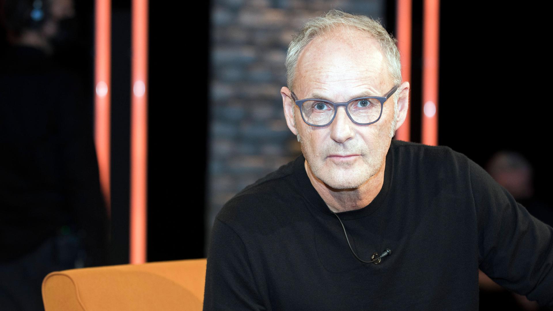 Moderator Reinhold Beckmann: Er sitzt in einem Fernsehstudio, trägt eine graue Brille und einen schwarzen Pullover.