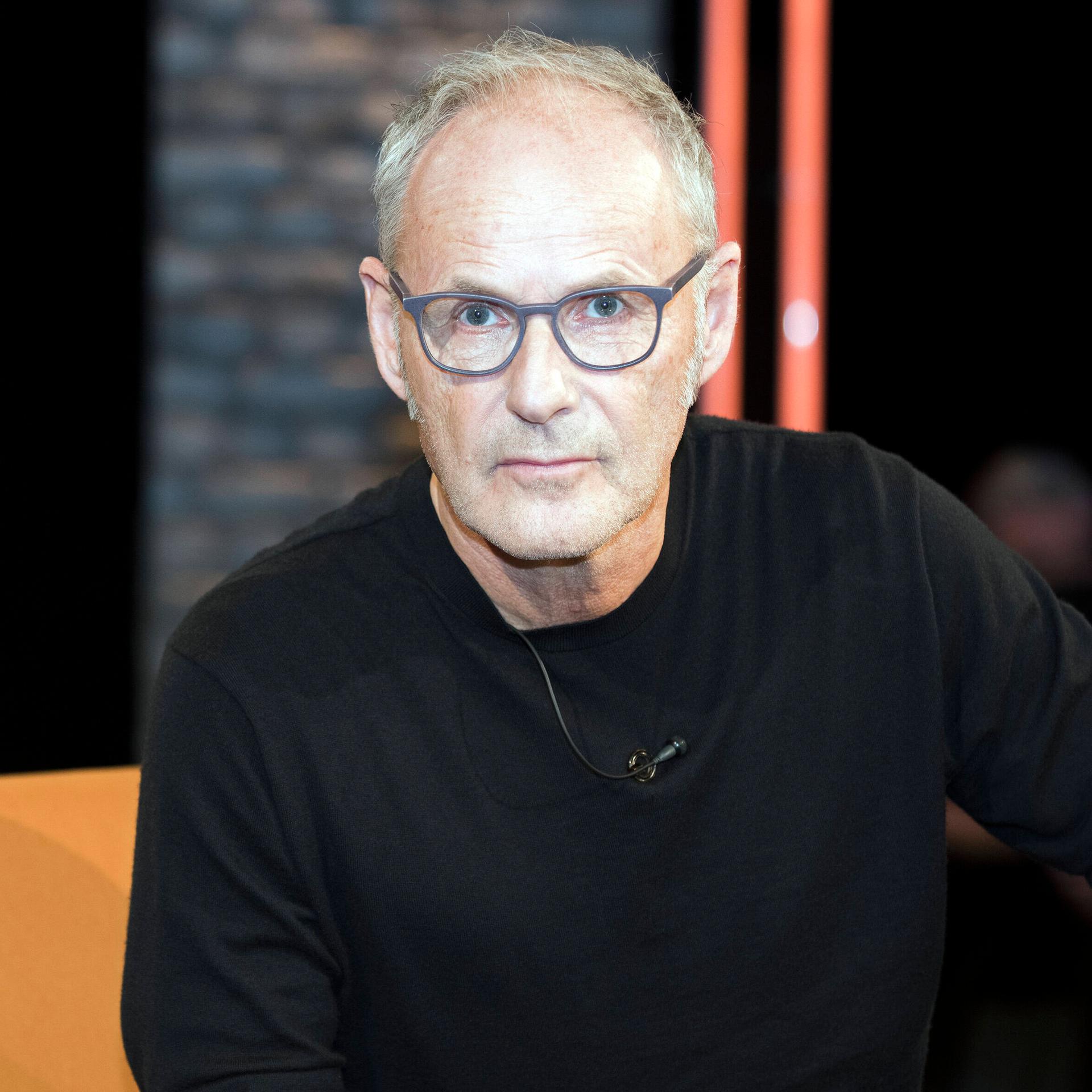 Moderator Reinhold Beckmann: Er sitzt in einem Fernsehstudio, trägt eine graue Brille und einen schwarzen Pullover.