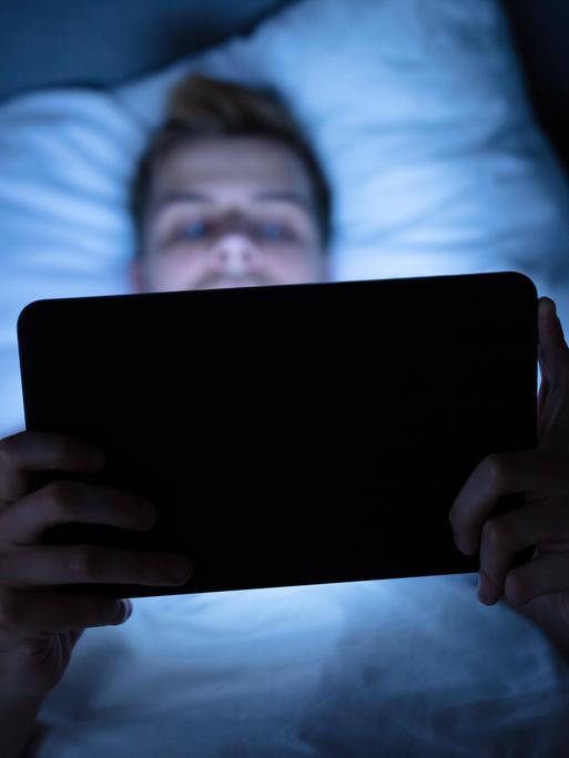 Das Foto eines Mannes, der auf dem Rücken im Bett liegt und ein Tablet in beiden Händen hält, auf das er schaut und das sein Gesicht blau beleuchtet. 