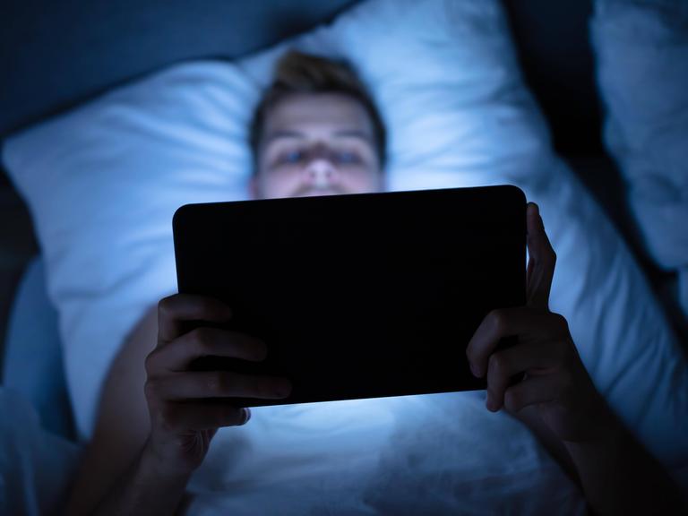 Das Foto eines Mannes, der auf dem Rücken im Bett liegt und ein Tablet in beiden Händen hält, auf das er schaut und das sein Gesicht blau beleuchtet. 