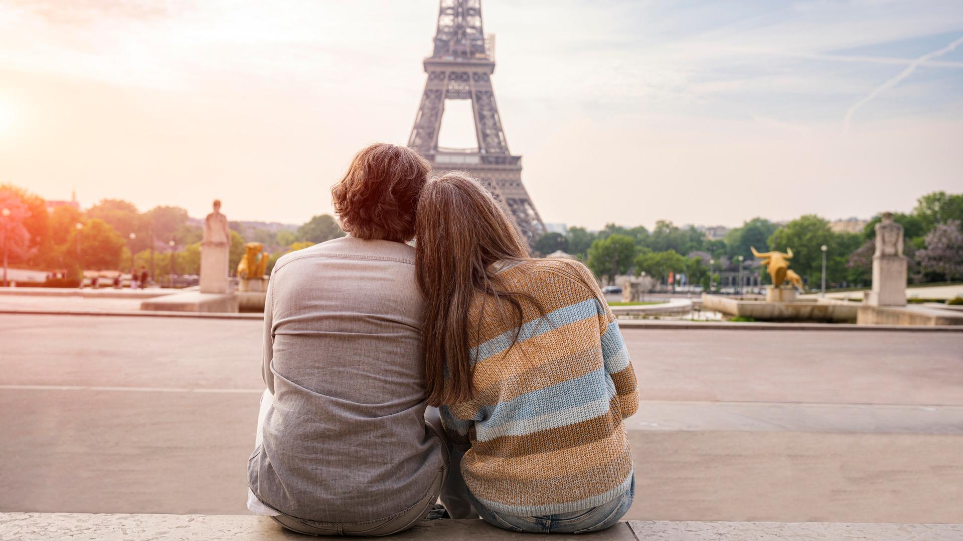 Ein Mann und eine Frau reiferen Alters sitzen eng aneinandergeschmiegt und betrachten den Eiffelturm in Paris. Sie sind in Rückenansicht zu sehen.