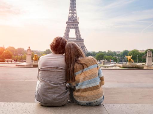 Ein Mann und eine Frau reiferen Alters sitzen eng aneinandergeschmiegt und betrachten den Eiffelturm in Paris. Sie sind in Rückenansicht zu sehen.