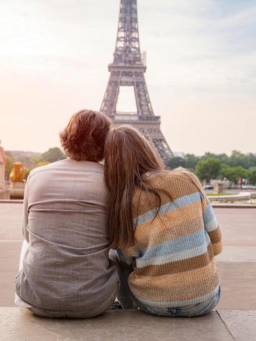 Ein Mann und eine Frau reiferen Alters sitzen eng aneinandergeschmiegt und betrachten den Eiffelturm in Paris. Sie sind in Rückenansicht zu sehen.