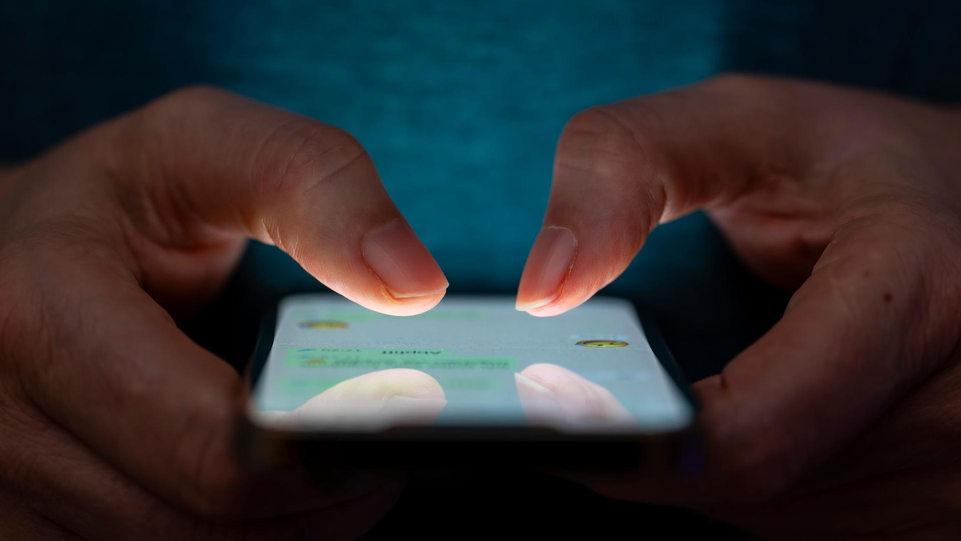 Man sieht zwei Hände vor einem dunklen Hintergrund. Sie tippen auf ein Handy. Man sieht zwei Hände vor einem dunklen Hintergrund. Sie tippen auf ein Handy.