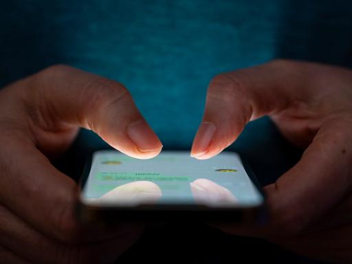 Man sieht zwei Hände vor einem dunklen Hintergrund. Sie tippen auf ein Handy. 