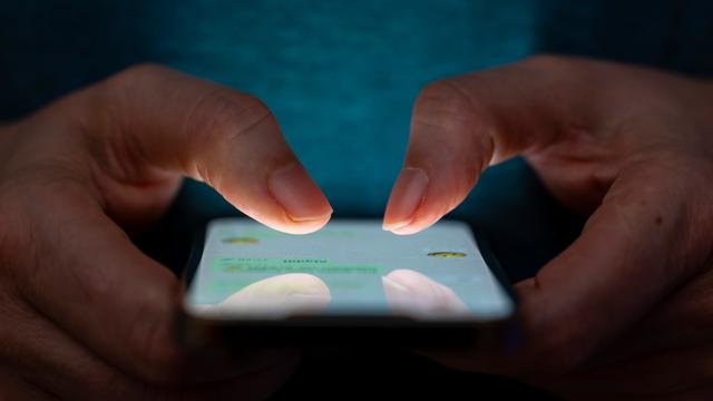 Man sieht zwei Hände vor einem dunklen Hintergrund. Sie tippen auf ein Handy. 