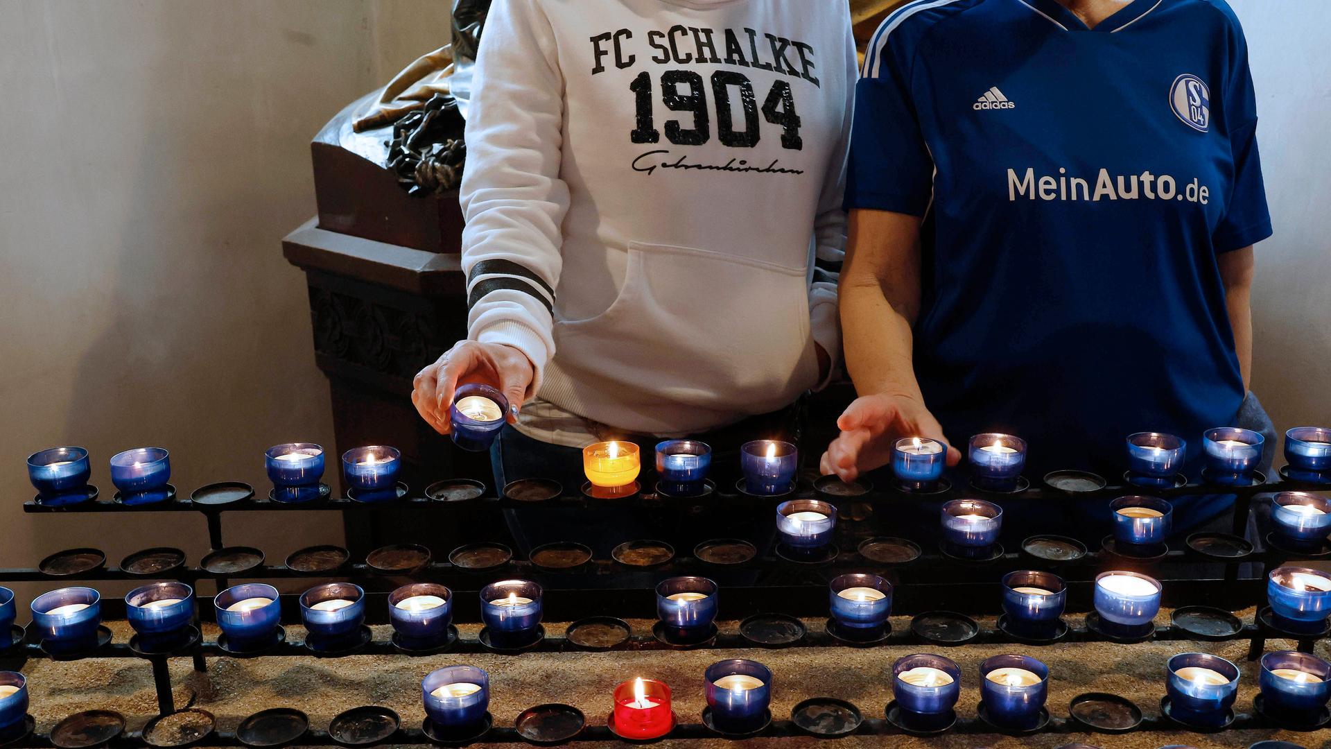 Zu sehen sind zwei Personen in Fankleidung von Schalke 04. Sie stehen vor einer Reihe an blau-weißen Teelichtern in einer Kirche. 