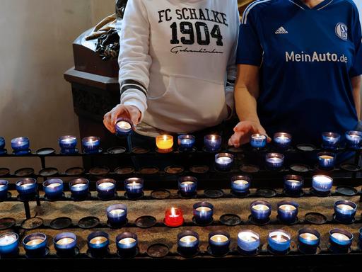 Zu sehen sind zwei Personen in Fankleidung von Schalke 04. Sie stehen vor einer Reihe an blau-weißen Teelichtern in einer Kirche. 